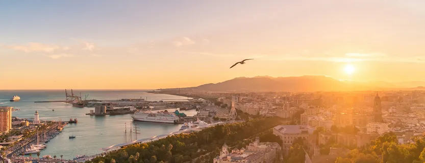 Aerial panoramic view of Málaga at sunset, with the Mediterranean Sea, the port, the Cathedral, and the mountains in the background.
